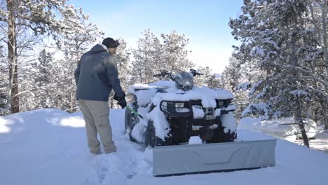 A-Caucasian-male-brushing-snow-off-of-an-ATV-or-4-wheeler-with-a-plow-to-clear-snow-from-a-mountain-driveway