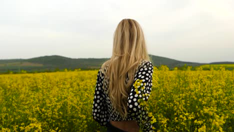Blond-woman-walking-in-field-of-yellow-rapeseed,-handheld-slow-motion