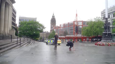 Two-young-men-skateboarding-through-Sheffield-city-centre,-England-on-a-rainy-overcast-day