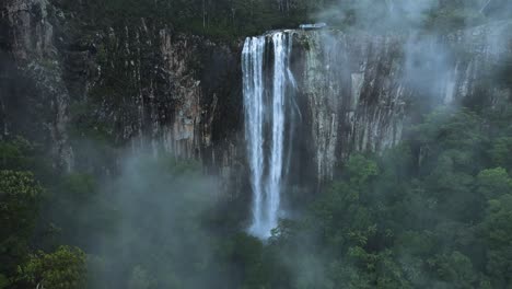 Unique-drone-view-through-suspended-mist-revealing-a-majestic-waterfall-spilling-down-a-lush-rainforest-mountain-scenery