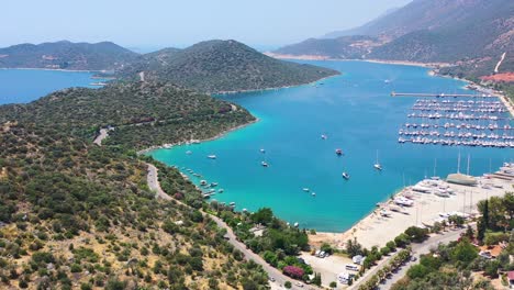 Aerial-drone-of-a-peninsula-full-of-green-hills-and-tropical-turquoise-blue-water-overlooking-the-Mediterranean-Sea-in-Kas-Turkey-on-a-sunny-summer-day-with-a-marina-and-docked-boats