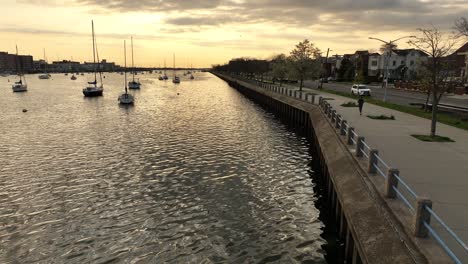 A-low-altitude-shot-over-Sheepshead-Bay-during-a-golden-sunrise-with-boats-anchored-and-a-jogger-running-on-the-paved-walkway-along-the-water