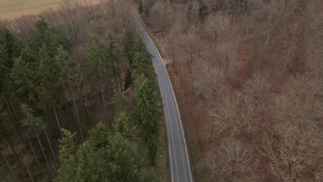 Bird's-eye-view-of-a-car-travelling-along-a-curved-countryside-highway-through-a-mixed-and-partially-bare-forest