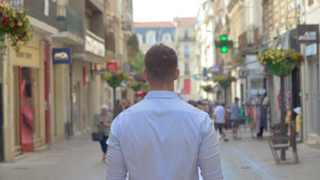 A-young-Caucasian-man-walking-with-his-back-to-a-pedestrian-street-in-the-south-of-France-with-shops-and-a-pharmacy-on-the-side