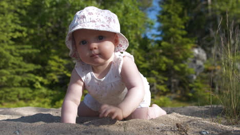 Baby-girl-with-big-blue-eyes-crawling-on-sand-looking-straight-at-camera