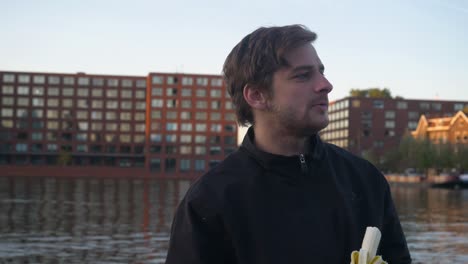 Primer-Plano-De-Un-Joven-Apuesto-Pelando-Y-Comiendo-Un-Plátano-Con-Un-Muelle-Urbano-En-El-Fondo,-Amsterdam