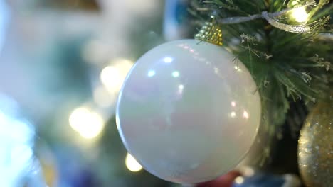 Nahaufnahme-Einer-Weißen-Glühbirne,-Die-An-Einem-Weihnachtsbaum-Hängt,-Während-Die-Lichter-Des-Baumes-Flackern-Und-Einen-Goldenen-Glanz-Erzeugen