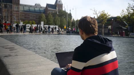 Handsome-young-man-sitting-on-the-banks-of-the-canal-in-front-of-Rijksmuseum-with-I-Amsterdam-sign-typing-a-text-on-laptop