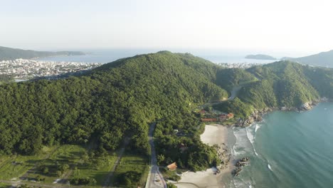 Cinematic-aerial-drone-video-of-jungle-mountains-with-a-road-revealing-a-city-behind-it,-Bombinhas,-Santa-Catarina,-Brazil