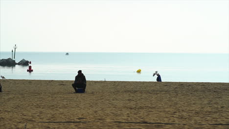 Slow-Motion-Silhouette-of-people-in-Barceloneta-beach,-sitting-and-walking-by,-with-birds-flying-around-next-to-a-rocky-pier