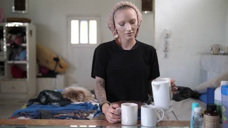 A-young,-white,-alternative-woman-with-a-unique-look-in-style-and-fashion-puts-two-tea-bags-in-two-mugs-and-pours-hot-boiling-water-from-the-boiler-into-them,-stands-up-and-walks-away