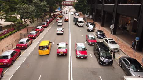 Traffic-passing-in-downtown-Hong-Kong-with-typical-Red-Taxis-parked-on-the-side-of-the-road,-Low-angle-aerial-view