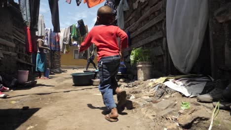Junge-Afrikanische-Kinder-Spielen-Rennend-Und-Gehen-Durch-Die-Slums-Eines-Armen-Dorfes,-Während-Sie-An-Ihrer-Mutter-Vorbeikommen,-Die-Wäsche-Wäscht-–-Folgende-Kamerafahrt-In-Zeitlupe-Mit-180-Bildern-Pro-Sekunde
