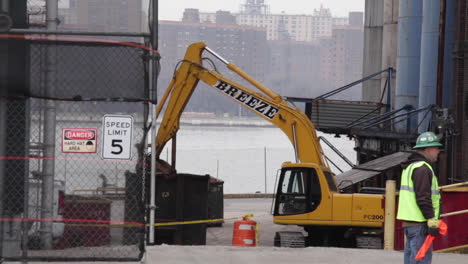 Excavator-Crane-Works-at-Construction-Site-on-East-River-in-Brooklyn,-Manhattan-in-Background