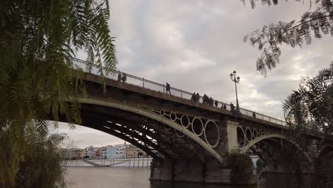 People-walk-across-landmark-Triana-Bridge,-Seville,-Spain,-cloudy-evening-sky