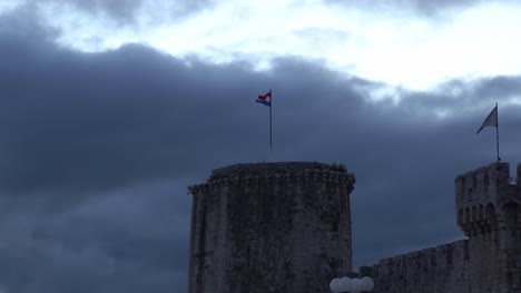 Tower-Kamerlengo,-in-the-town-of-Trogir-in-Croatia,-at-sunset-with-a-long-Croatian-flag-and-clouds-drifting-by