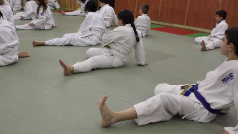 Estudiantes-De-Karate-En-Clase-Se-Estiran-Para-Calentar-Para-La-Práctica,-Uniformes-Blancos