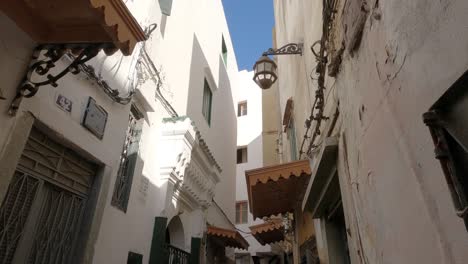 Picturesque-Narrow-street-with-tiled-door-porticos,-People-walking-by-Tangier-shops,-Tilt-down-shot