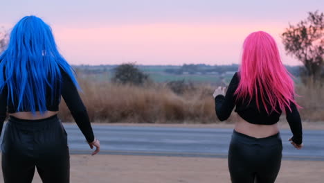 Two-women-with-pink-and-blue-wigs-running-towards-a-busy-road-in-the-sunset