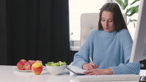 Young-woman-writing-something-in-notebook-while-sitting-at-office-desk-There-are-red-apples-and-healthy-salad-bowl-on-the-desk