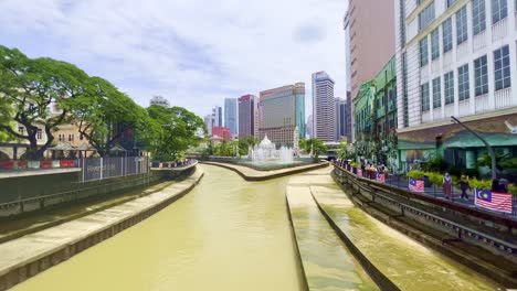 High-angle-shot-over-river-flowing-with-Sultan-Abdul-Samad-Jamek-mosque-visible-in-distance-along-with-tall-buildings-in-Kuala-Lumpur,Malaysia-on-a-sunny-day
