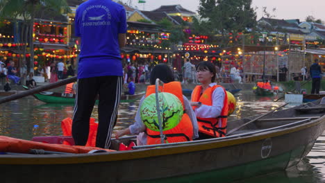 Tourists-taking-lantern-boat-ride-on-Hoai-River-in-traditional-Sampan-boat-in-ancient-town-of-Hoi-An-at-evening-with-city-view-in-background,-Vietnam
