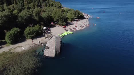 Aerial-shot-moving-forward-of-a-beach-full-of-people-with-an-abandoned-boat-in-the-alpine-glacier-lake-surrounded-by-forest-in-Bariloche,-Colonia-Suiza,-Patagonia-Argentina