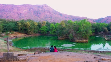 beautiful-couple-sitting-on-rock,-a-mountain-in-the-middle-of-forest-area-and-river-view,-Drone-Camera-moving-forward-overhead-to-couple,-couple-relaxing-on-naturel-beauty