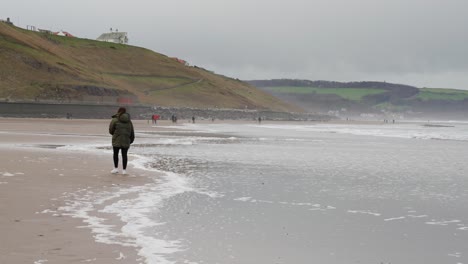 Brunette-girl-walks-along-a-quiet-english-beach-in-the-distance-on-a-cold-windy-afternoon-as-the-tide-comes-in-and-waves-approach-her-to-splash-her