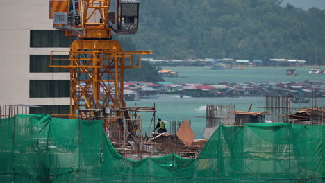 Worker-People-Working-at-Apartment-Building-Construction-Site-with-Crane-on-Top,-Kota-Kinabalu-City-Water-Villages-by-Gaya-Island-in-Background---close-up-slow-motion