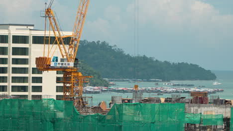 Aerial-view-of-People-Working-at-Apartment-Building-Construction-Site-with-Crane-on-Top,-Kota-Kinabalu-City-Water-Villages-by-Gaya-Island-in-Background