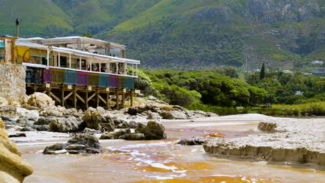 Breached-lagoon-with-tannin-rich-water-runs-toward-ocean-past-Milk-On-The-Beach-restaurant,-Onrus,-South-Africa