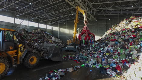 Digger-and-crane-loading-heap-of-trash-in-recycling-yard-hall,aerial-view