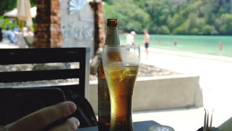 Cerveza-Fría-En-Una-Mesa-Con-Una-Playa-De-Arena-Blanca-Al-Fondo