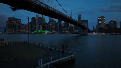 Eine-Luftaufnahme-Von-Einem-Brooklyn-Park-Unter-Der-Brooklyn-Bridge-Mit-Dem-Freedom-Tower-In-Lower-Manhattan-Im-Hintergrund-Vor-Einem-Wolkigen-Sonnenaufgang