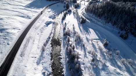 Aerial-reveal-of-snow-covered-mountain-landscape-with-a-river,-forests-a-road-and-cars-passing-and-villages-on-a-sunny-day-in-Pontresina,-Switzerland