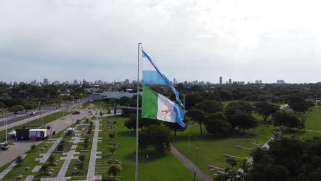Aerial-orbit-of-Argentina-flag-reveals-park,-modern-city,-and-roadway