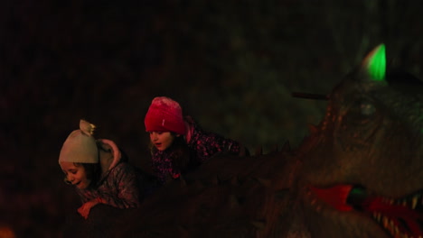 Two-curious-little-girls-having-fun-riding-a-dinosaur-ride-at-an-amusement-parka-at-night-time