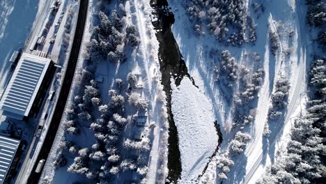 Aerial-top-down-view-of-snow-covered-landscape-with-a-river,-forests-a-road-and-cars-passing-on-a-sunny-day-in-Pontresina,-Switzerland