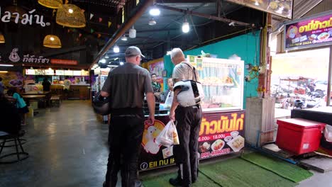 2-tourists-men-buying-food-in-an-indoor-food-market-while-holding-motorbike-helmets