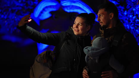 Young-couple-with-baby-taking-a-selfie-in-an-amusement-park-lit-up-at-night