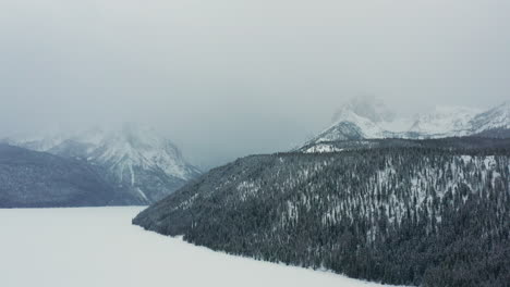 Aerial-moving-over-frozen-lake-and-forest-towards-large-mountains-in-fog