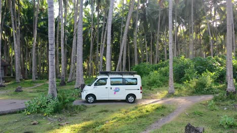 white-camper-van-driving-through-a-tropical-coconut-tree-field-at-sunset-in-Bali,-aerial