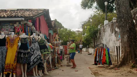 Small-street-clothing-shop-full-of-colorfull-dresses-with-old-woman-with-mask-on-a-cloudy-day