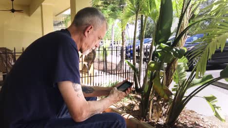 Elderly-man-sitting-using-his-mobile-device-in-a-tropical-setting