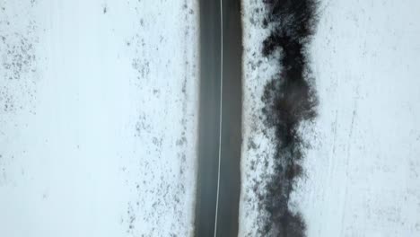 Empty-road-in-winter,-landscape-covered-in-snow,-birds-eye-drone-shot,-black-car-driving-on-the-road,-eerie-feel-and-anticipation