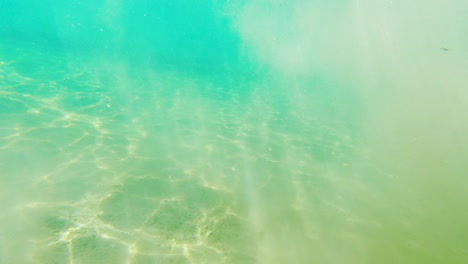 Swimming-in-Rays-of-Sunlight-and-Floating-Sand-in-Shallow-Teal-Lake,-Wide-POV-Underwater-Dolly-In