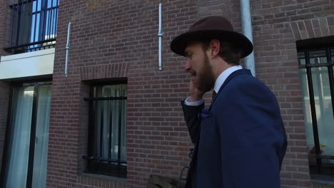 An-elegant-man-in-dandy-style-with-blue-suit-and-beard-is-talking-on-a-cellphone-while-walking-against-a-brick-wall-in-Amsterdam