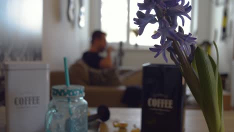 Im-Hintergrund-Verschwommener-Mann,-Der-Morgens-Auf-Dem-Sofa-Sitzt-Und-Kaffee-Trinkt,-Mit-Einer-Violetten-Blume-Im-Vordergrund