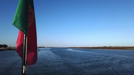Portuguese-flag-hangs-in-foreground-on-ferry-navigating-Ria-Formosa-near-Olhao,-Portugal,-close-up